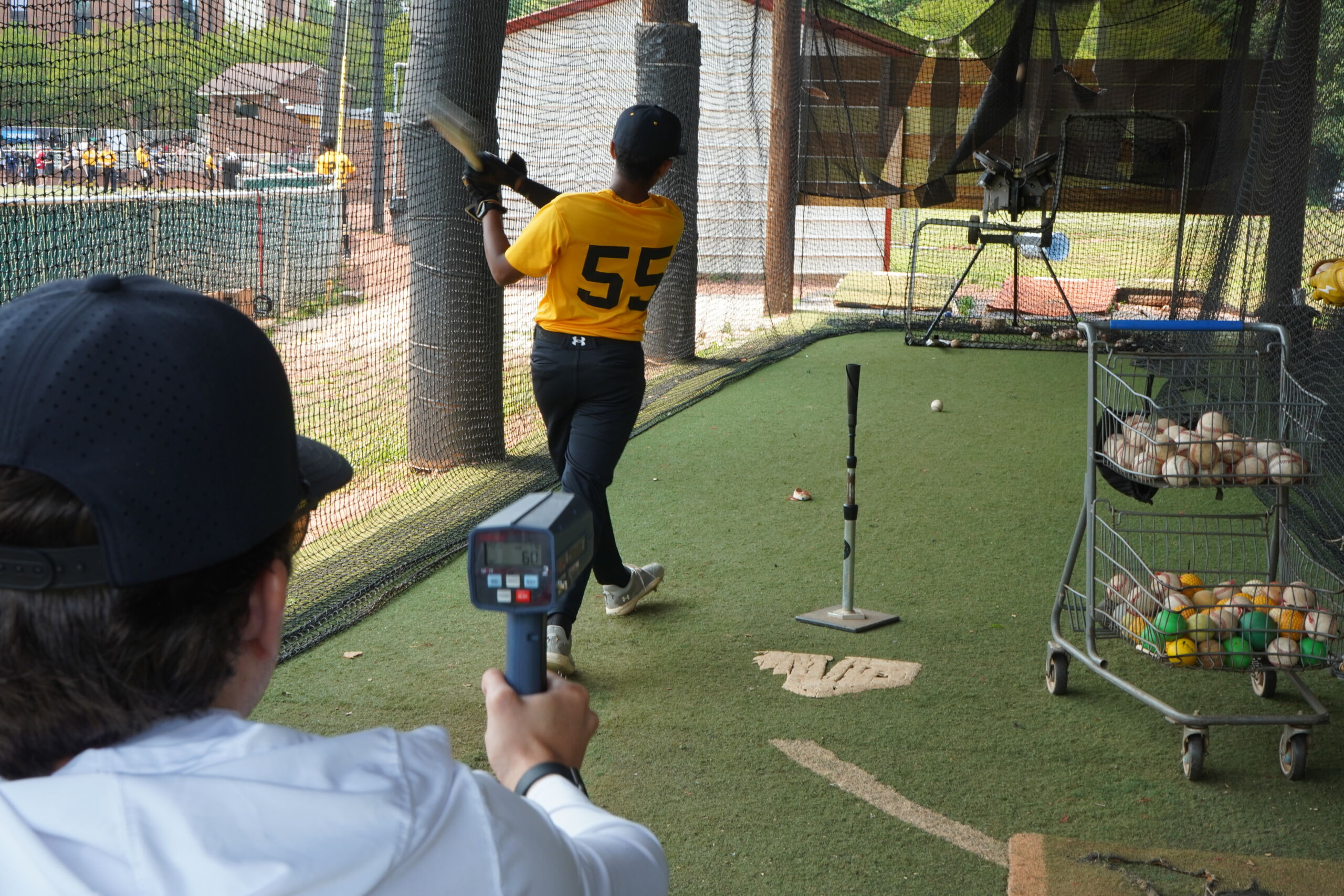 Baseball player hitting off a tee during Committed Baseball Showcase batting evaluation as coach records exit velocity with radar gun.