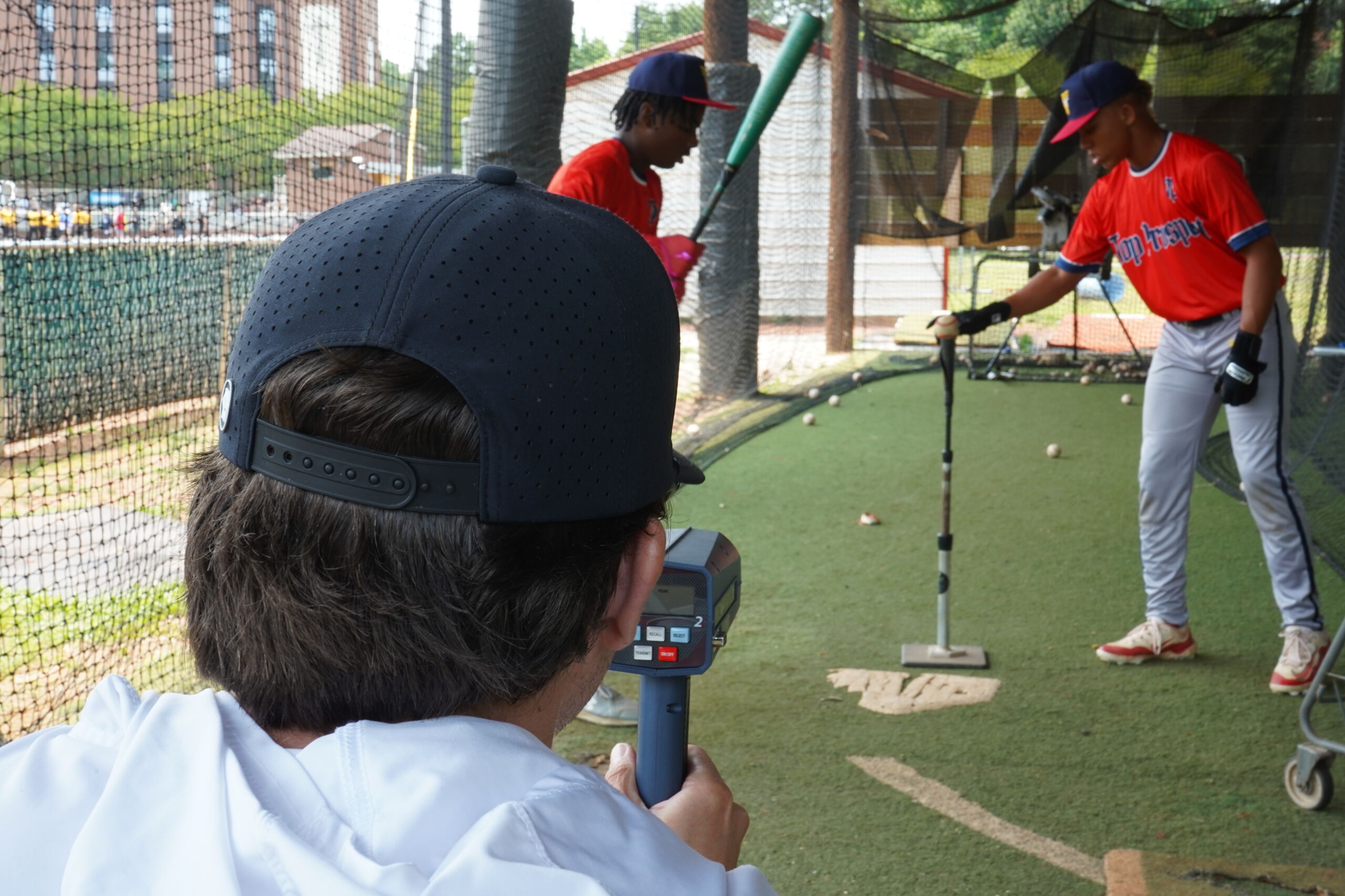 Baseball player hitting off a tee during Committed Baseball Showcase batting evaluation as coach records exit velocity with radar gun.