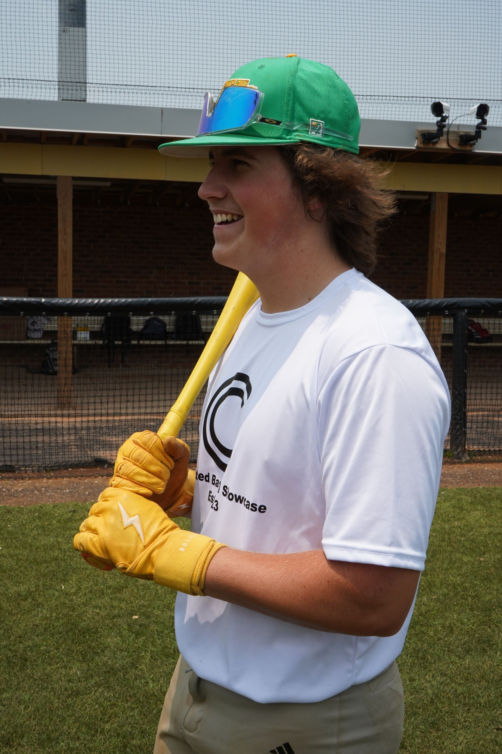 Smiling baseball prospect holds bat during Committed Baseball Showcase, wearing branded shirt and preparing for hitting session in front of college coaches from NC and VA.