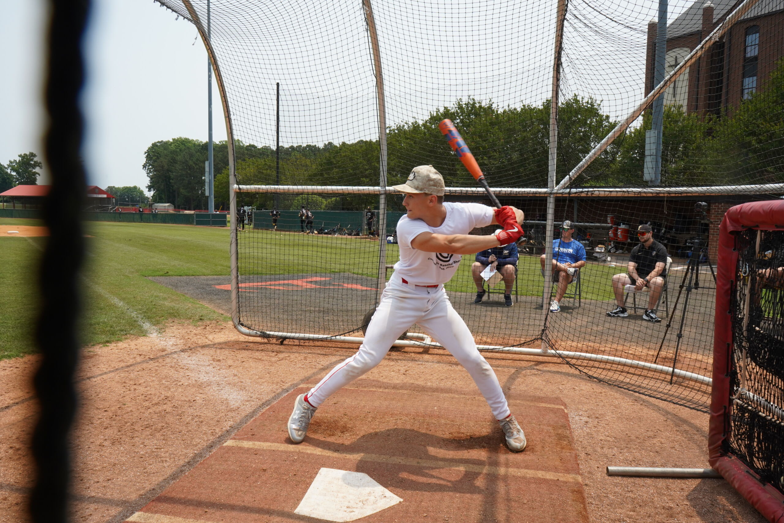 High school hitter takes batting practice in front of college coaches during Committed Baseball Showcase at outdoor field.