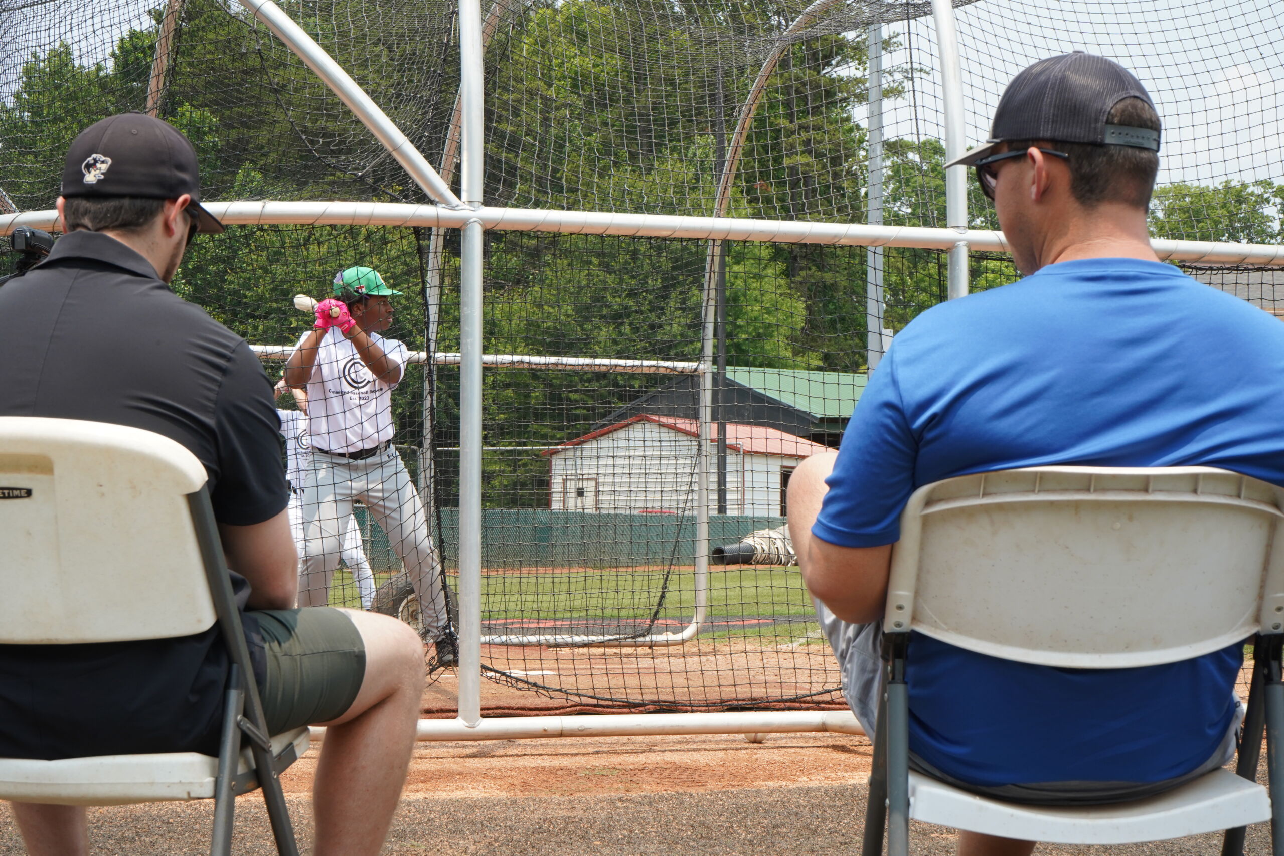 College coaches scout hitter during live batting practice at Committed Baseball Showcase with player in cage and evaluators seated behind net.